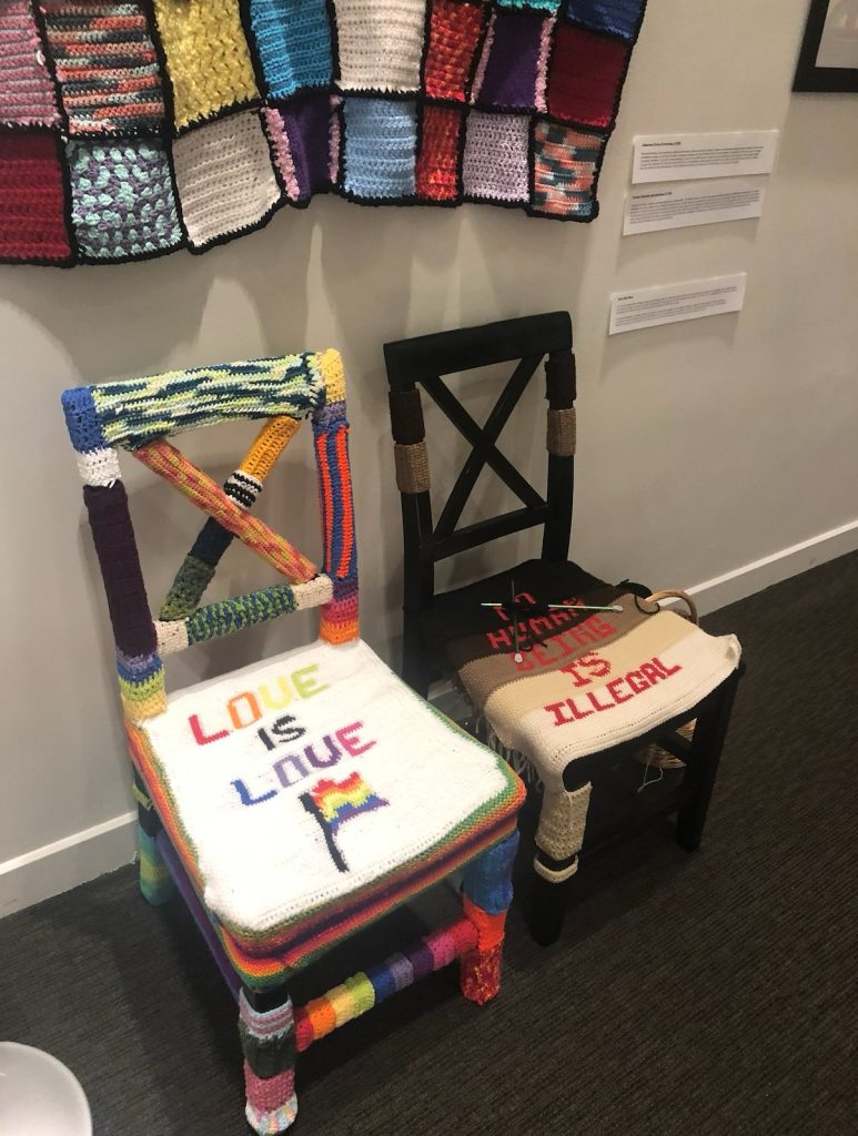 Two crocheted chairs, one reading Love is love and one reading No human being is illegal, in the installation Sit in my place