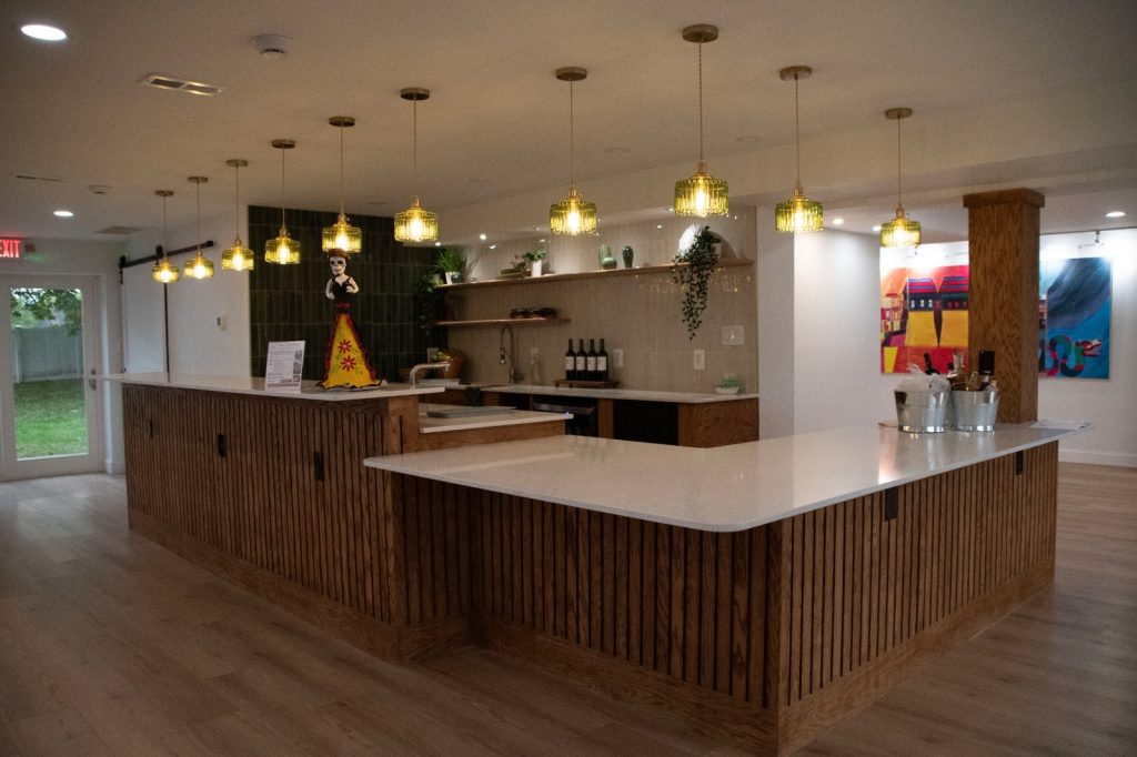 The new bar and gathering space with wood-slat counters, white countertops and a row of green glass pendant lamps