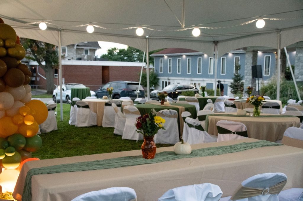 Round tables with white chair covers, green runners, flowers and small white pumpkins under a white tent