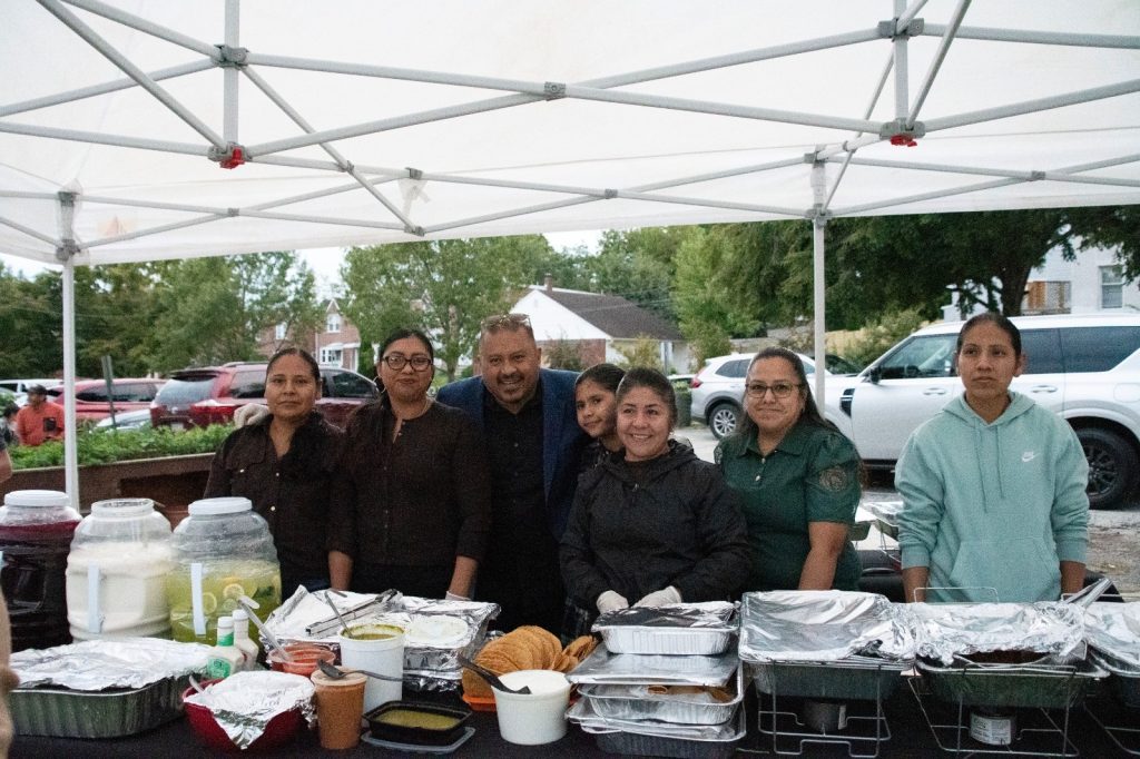 Seven members of the food committee smiling behind a long table of covered trays and aguas frescas